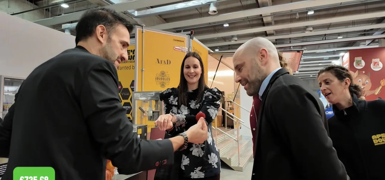 Sergio Starman esegue un gioco con le palline rosse davanti a uno stand in fiera, i visitatori sorridono